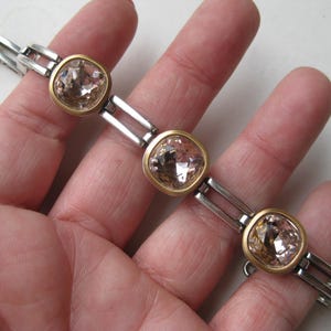 May include: A bracelet featuring three square-shaped, faceted light champagne-colored crystals set in gold-toned bezels. The crystals are connected by silver-toned rectangular metal links, creating a structured chain design with a metallic finish.
