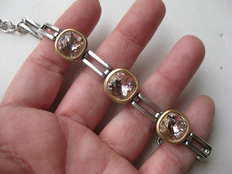 May include: A bracelet featuring three square-shaped, faceted light champagne-colored crystals set in gold-toned bezels. The crystals are connected by silver-toned rectangular metal links, creating a structured chain design with a metallic finish.