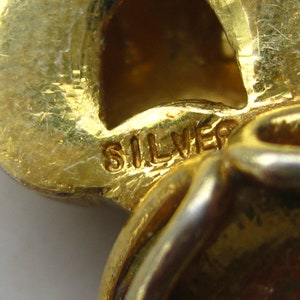 May include: A gold-toned metal jewelry piece featuring a textured surface and a stamped mark that reads SILVER. The object has a curved, organic shape with a hollow section and a polished finish, showing clear signs of wear and metallic texture.