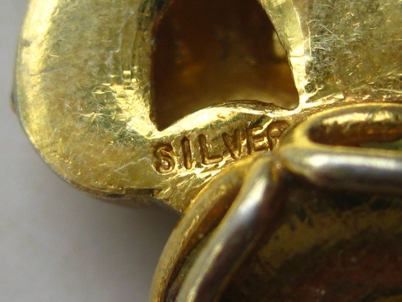 May include: A gold-toned metal jewelry piece featuring a textured surface and a stamped mark that reads SILVER. The object has a curved, organic shape with a hollow section and a polished finish, showing clear signs of wear and metallic texture.