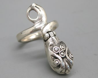 May include: A silver-toned ring with an ornate, textured design. The ring features a loop and a detailed, pod-like shape with circular and curved embellishments. The ring is set against a gray background.