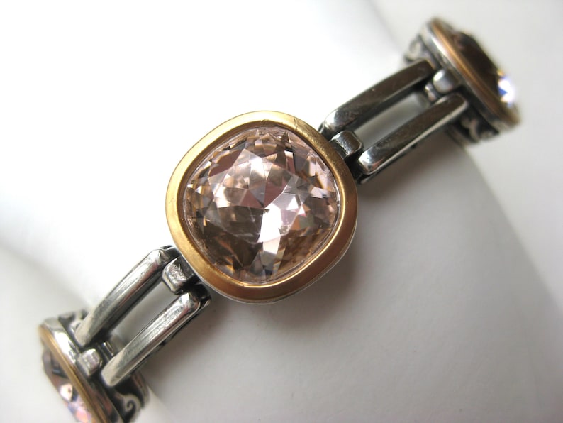 May include: A link bracelet featuring square-shaped, faceted light pink stones set in gold-toned bezels. The stones are connected by polished silver-toned metal links with decorative scrollwork details on the sides of each stone setting.