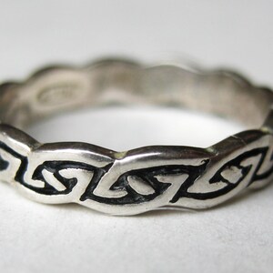 May include: A silver-toned metal ring featuring a continuous, raised, wavy band design with recessed black detailing that creates a repeating geometric pattern around the entire circumference of the circular jewelry piece.