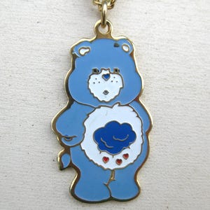 May include: A gold-toned metal pendant shaped like a blue bear character with white facial details and a white belly patch featuring a dark blue cloud icon with three small red hearts, suspended from a gold-toned metal chain link.