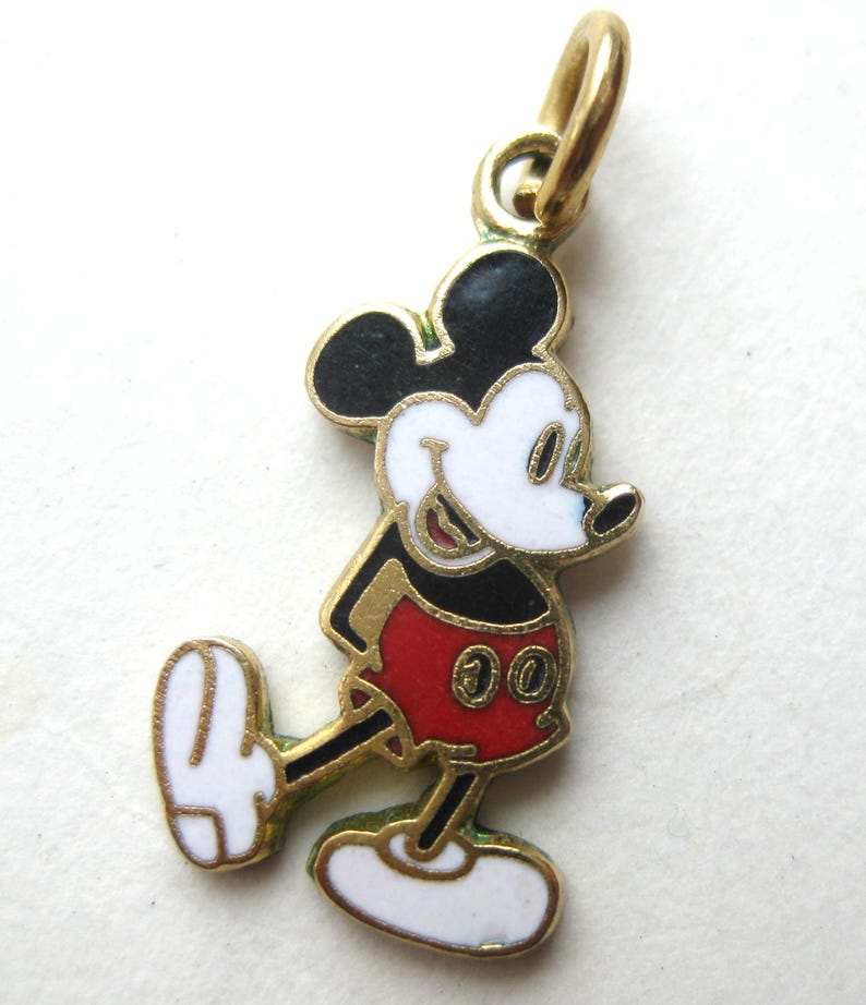 Puede incluir: Un charm de Mickey Mouse dorado con orejas negras, cara blanca y pantalones cortos rojos. El charm tiene un peque&ntilde;o lazo en la parte superior para sujetarlo. El personaje de dibujos animados cl&aacute;sico se representa caminando.