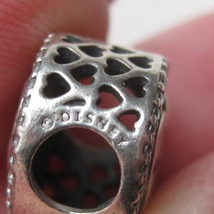 May include: A silver-toned metal charm bead featuring a repeating cut-out heart pattern across its surface. The side of the bead displays the text DISNEY engraved in capital letters, accompanied by a copyright symbol, surrounding a central hole.