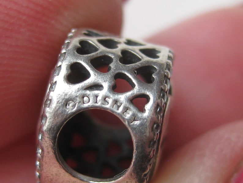 May include: A silver-toned metal charm bead featuring a repeating cut-out heart pattern across its surface. The side of the bead displays the text DISNEY engraved in capital letters, accompanied by a copyright symbol, surrounding a central hole.