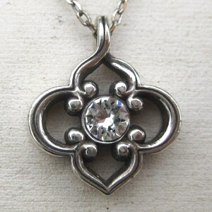 May include: A silver-toned metal pendant necklace featuring a quatrefoil-shaped frame with scrolled edges and a single, round, clear faceted gemstone set in the center. The pendant hangs from a thin, matching silver-toned metal chain link necklace.
