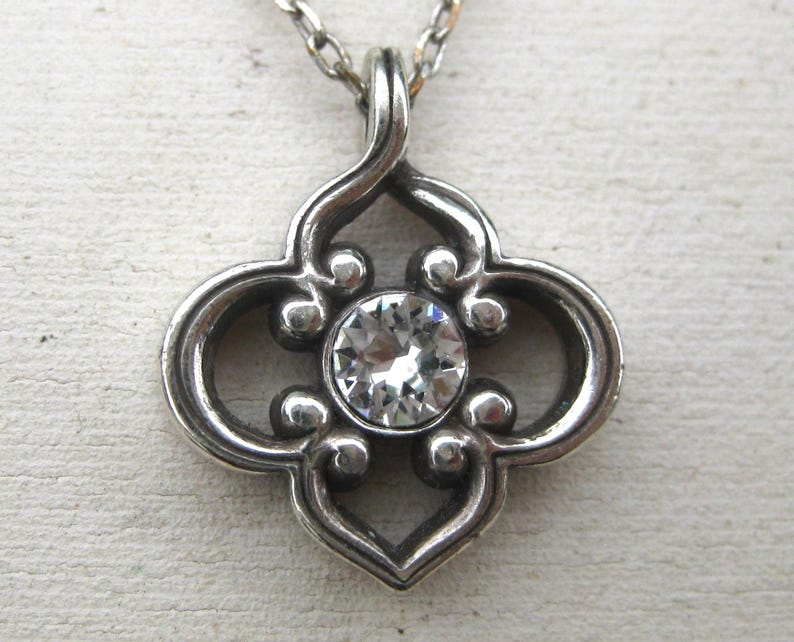 May include: A silver-toned metal pendant necklace featuring a quatrefoil-shaped frame with scrolled edges and a single, round, clear faceted gemstone set in the center. The pendant hangs from a thin, matching silver-toned metal chain link necklace.