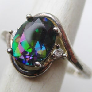 May include: A silver ring featuring an oval-cut, iridescent gemstone with hues of green, purple, and blue. The ring has a swirling design and is accented with two small, clear gemstones. The ring is a piece of jewelry.