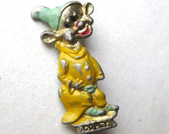 May include: A vintage metal pin featuring a cartoon character wearing a yellow outfit and a light green hat. The character has a red mouth and is standing on a base with the name Dopey embossed in silver lettering. The metal shows signs of wear.