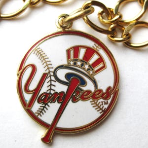 Op de afbeelding: Een goudkleurige ketting met een ronde hanger met het logo van de New York Yankees. De hanger heeft een witte achtergrond met een rood en blauw ontwerp van een honkbalknuppel en een hoed. Het woord "Yankees" is rood geschreven.