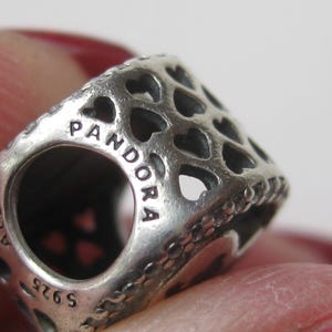 May include: A silver charm bead featuring a repeating cut-out heart pattern across its surface. The metal is engraved with the text PANDORA, ALE, and S925. The bead has a cylindrical shape with a central opening for a bracelet or necklace chain.