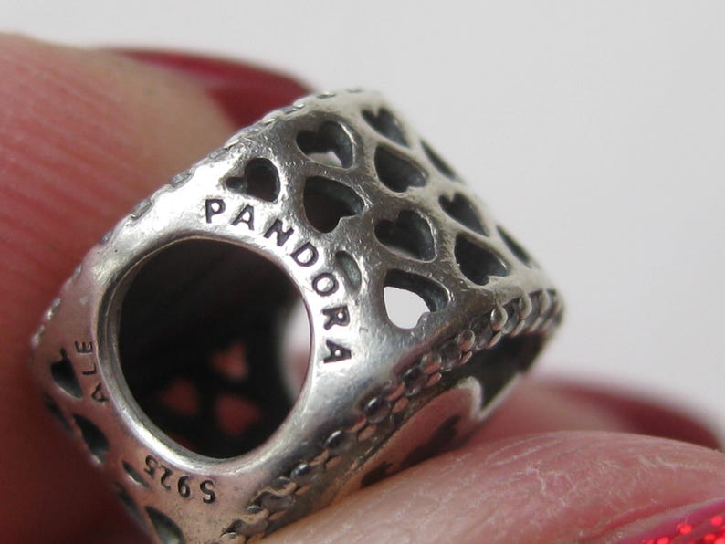 May include: A silver charm bead featuring a repeating cut-out heart pattern across its surface. The metal is engraved with the text PANDORA, ALE, and S925. The bead has a cylindrical shape with a central opening for a bracelet or necklace chain.