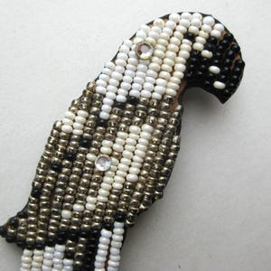 May include: A beaded parrot pin with a brown backing. The parrot is made of white, black, and silver beads. Two clear rhinestones are placed on the parrot's body. The parrot's beak is black.