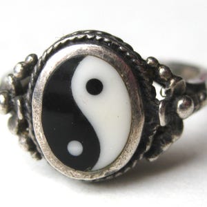May include: A silver ring featuring an oval-shaped inlay with a black and white yin-yang symbol. The metal band has a textured, rope-like border surrounding the central inlay and decorative floral-inspired metalwork on the sides of the setting.