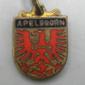 May include: A brass-toned metal charm shaped like a shield, featuring the text APELDOORN in a black rectangular bar at the top. Below the text, a red eagle is depicted with a key held in its talons, set against a textured brass background.