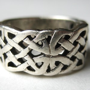 May include: A silver-colored ring with a Celtic knot design. The ring features an intricate, interwoven pattern that wraps around the entire band. The metal has a slightly aged appearance, with visible details in the knotwork.