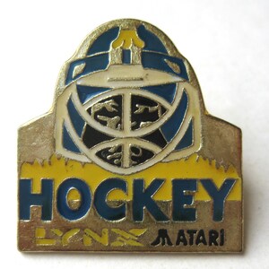 Könnte beinhalten: Vintage Hockey-Anstecknadel mit goldfarbenem Rand. Die Anstecknadel zeigt ein Hockey-Torwartmasken-Design in Blau, Schwarz und Weiß, mit dem Wort "HOCKEY" in Blau und Gelb. Der Text "LYNX ATARI" befindet sich unten.