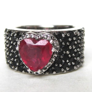 May include: A wide silver band ring featuring a large, faceted, heart-shaped red gemstone at the center. The heart is surrounded by a border of small clear stones, while the rest of the band is densely encrusted with numerous small black stones.