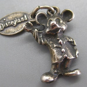 May include: A silver-toned Mickey Mouse charm with the word "Disneyland" on a tag. The charm depicts Mickey Mouse in a classic pose, with his arm outstretched. The charm is detailed and has a vintage look.
