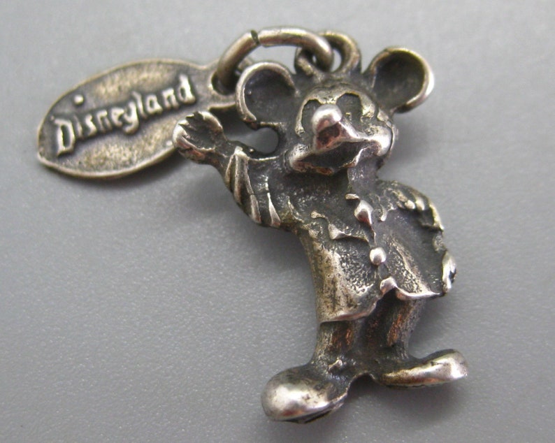 May include: A silver-toned Mickey Mouse charm with the word "Disneyland" on a tag. The charm depicts Mickey Mouse in a classic pose, with his arm outstretched. The charm is detailed and has a vintage look.
