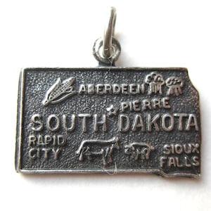 May include: A silver-toned charm in the shape of South Dakota. The charm features embossed images of a corn stalk, cattle, and a pig, with the state name and city names: Aberdeen, Pierre, Rapid City, and Sioux Falls.