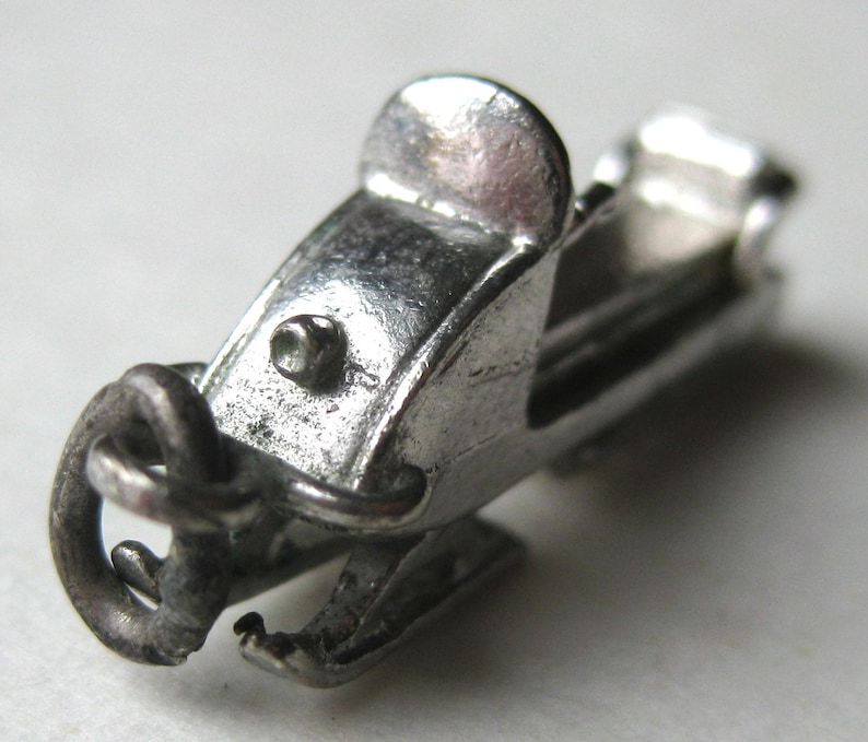 Może przedstawiać: Mały, srebrny charms w kształcie skutera śnieżnego z misternymi detalami. Charms posiada siedzenie, kierownicę i pętlę do przymocowania do naszyjnika lub bransoletki. Projekt skutera śnieżnego jest bardzo szczeg&oacute;łowy, z widocznymi gąsienicami i teksturowaną powierzchnią.