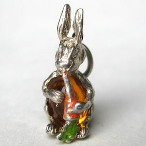 May include: A silver-toned bunny charm holding a carrot. The bunny has detailed features, with a carrot in orange, red, and green. The charm is likely made of metal and has a loop for attaching to a bracelet or necklace.