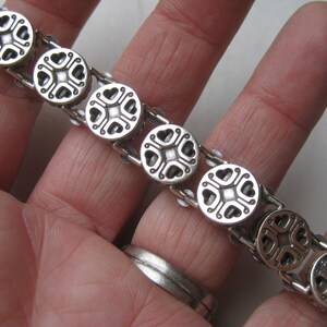 Puede incluir: Una pulsera de metal plateado compuesta por una serie de eslabones circulares. Cada eslab&oacute;n circular presenta un dise&ntilde;o sim&eacute;trico y hundido con cuatro recortes en forma de coraz&oacute;n dispuestos alrededor de un punto cuadrado central.