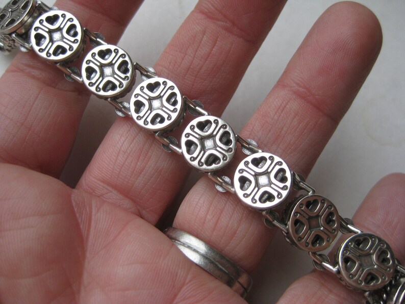 Puede incluir: Una pulsera de metal plateado compuesta por una serie de eslabones circulares. Cada eslab&oacute;n circular presenta un dise&ntilde;o sim&eacute;trico y hundido con cuatro recortes en forma de coraz&oacute;n dispuestos alrededor de un punto cuadrado central.