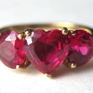 May include: A gold ring featuring three heart-shaped, faceted, deep red gemstones. The gemstones are set in a gold band with small gold prongs holding each stone in place. The ring is a close-up shot against a white background.