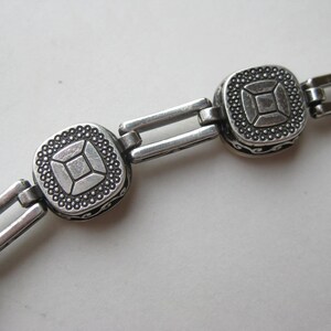 May include: A silver-toned metal bracelet featuring a series of square links with a central geometric pattern, surrounded by a beaded border. The links are connected by double rectangular bars, and the sides of the square components show black scrollwork.