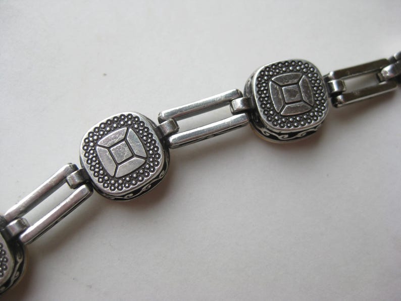 May include: A silver-toned metal bracelet featuring a series of square links with a central geometric pattern, surrounded by a beaded border. The links are connected by double rectangular bars, and the sides of the square components show black scrollwork.
