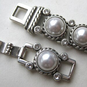 Puede incluir: Una pulsera de metal plateado con una serie de perlas blancas redondas de imitaci&oacute;n engastadas en bordes de estilo cuerda trenzada. Cada perla est&aacute; flanqueada por peque&ntilde;os cristales transparentes de corte redondo y un cierre de caja rectangular.