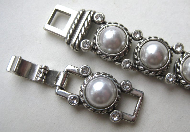Puede incluir: Una pulsera de metal plateado con una serie de perlas blancas redondas de imitaci&oacute;n engastadas en bordes de estilo cuerda trenzada. Cada perla est&aacute; flanqueada por peque&ntilde;os cristales transparentes de corte redondo y un cierre de caja rectangular.