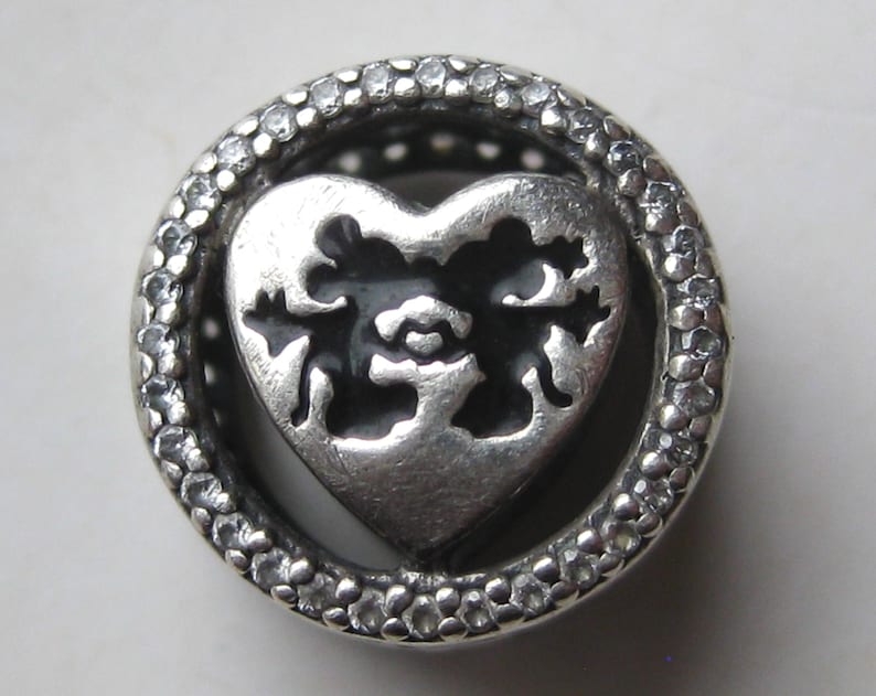 May include: A circular silver charm bead featuring a central heart cutout with a black enamel background. The outer rim of the bead is encrusted with small, clear, sparkling stones set in a textured, metallic frame surrounding the heart.
