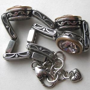 May include: A silver-toned link bracelet featuring three circular settings with clear faceted stones rimmed in gold. The links have an oxidized finish with embossed scrollwork patterns. The bracelet includes a lobster claw clasp and a small heart-shaped charm.