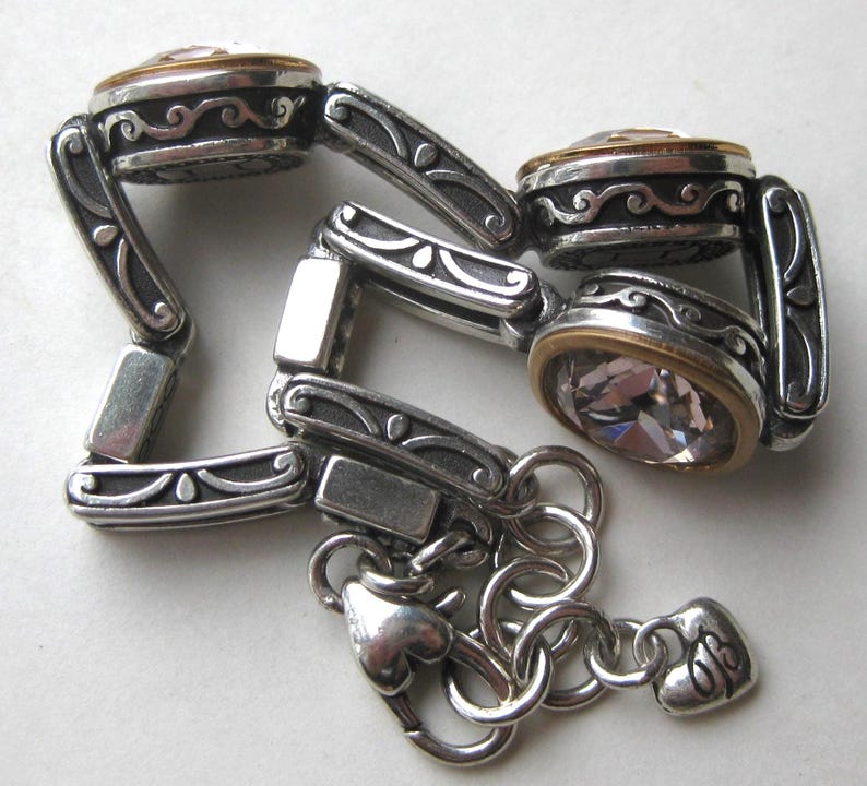 May include: A silver-toned link bracelet featuring three circular settings with clear faceted stones rimmed in gold. The links have an oxidized finish with embossed scrollwork patterns. The bracelet includes a lobster claw clasp and a small heart-shaped charm.