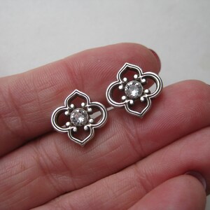 May include: A pair of silver-toned stud earrings featuring a four-lobed, openwork quatrefoil design. Each earring is centered with a single, clear, round-cut faceted stone set within the metal frame, creating a symmetrical and detailed appearance.
