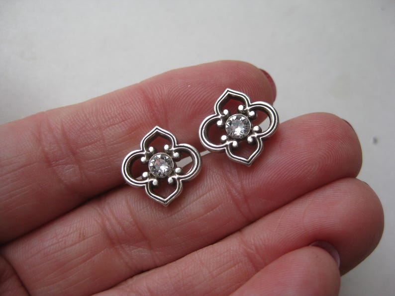 May include: A pair of silver-toned stud earrings featuring a four-lobed, openwork quatrefoil design. Each earring is centered with a single, clear, round-cut faceted stone set within the metal frame, creating a symmetrical and detailed appearance.