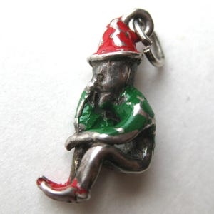 May include: A silver-toned metal charm shaped like a seated figure wearing a pointed hat. The figure features red enamel on the hat and shoes, and green enamel on the torso. A small silver jump ring is attached to the top of the pointed hat.