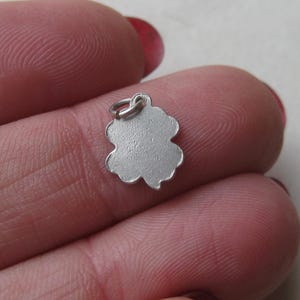 K&ouml;nnte beinhalten: Ein kleiner, silberfarbener Kleeblatt-Anh&auml;nger mit strukturierter Oberfl&auml;che und einer Schlaufe zum Befestigen an einer Halskette oder einem Armband. Der Anh&auml;nger wird zwischen den Fingern gehalten und zeigt sein filigranes Design.