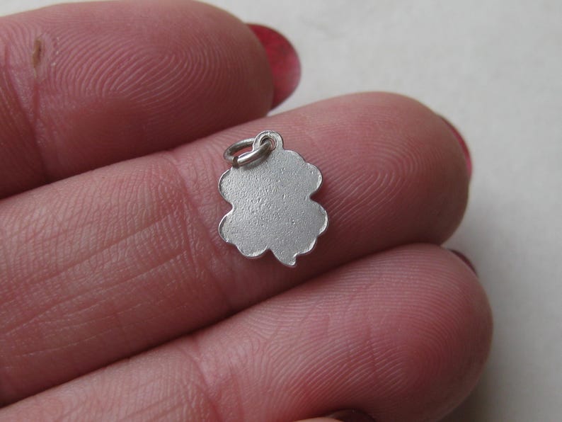 K&ouml;nnte beinhalten: Ein kleiner, silberfarbener Kleeblatt-Anh&auml;nger mit strukturierter Oberfl&auml;che und einer Schlaufe zum Befestigen an einer Halskette oder einem Armband. Der Anh&auml;nger wird zwischen den Fingern gehalten und zeigt sein filigranes Design.