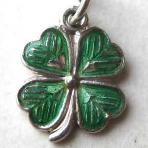 K&ouml;nnte beinhalten: Ein silberner Anh&auml;nger in Form eines vierbl&auml;ttrigen Kleeblatts mit gr&uuml;ner Emaille. Die Bl&auml;tter des Kleeblatts sind mit silbernen Konturen und Adern versehen. Der Anh&auml;nger h&auml;ngt an einer silbernen Schlaufe.