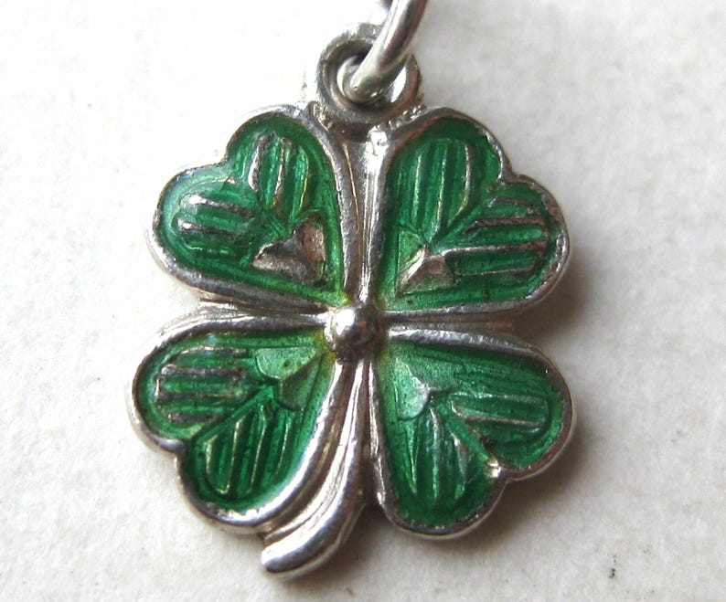 K&ouml;nnte beinhalten: Ein silberner Anh&auml;nger in Form eines vierbl&auml;ttrigen Kleeblatts mit gr&uuml;ner Emaille. Die Bl&auml;tter des Kleeblatts sind mit silbernen Konturen und Adern versehen. Der Anh&auml;nger h&auml;ngt an einer silbernen Schlaufe.