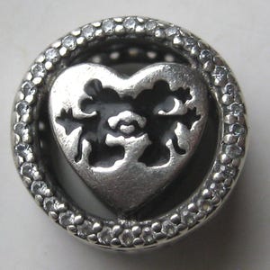 May include: A circular silver charm bead featuring a central heart cutout design with black enamel detailing. The outer ring of the bead is encrusted with small, clear, sparkling stones, creating a textured, metallic, and reflective appearance.