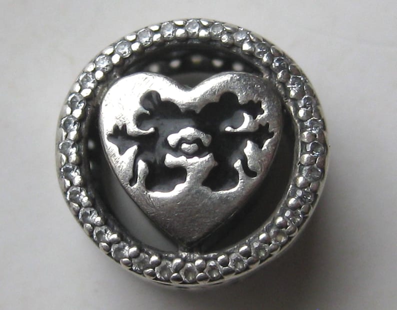 May include: A circular silver charm bead featuring a central heart cutout design with black enamel detailing. The outer ring of the bead is encrusted with small, clear, sparkling stones, creating a textured, metallic, and reflective appearance.