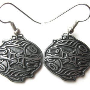 May include: A pair of silver-toned earrings with a detailed, engraved design. Each earring features a stylized, symmetrical motif with curved lines and shapes, suspended from a simple hook.