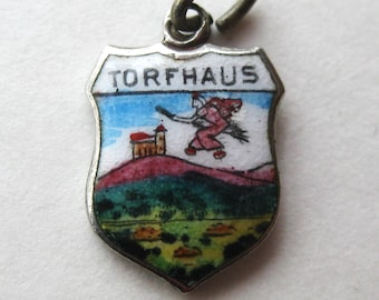 May include: A shield-shaped metal charm featuring a white enamel top section with the black text TORFHAUS. Below, a colorful scene depicts a figure on a broomstick flying over a small building on a hill, with green and yellow landscape details.